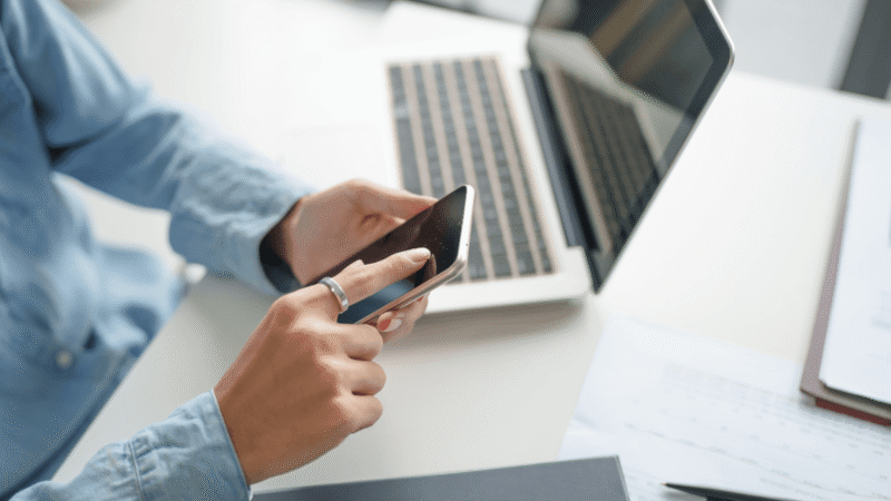 Businessperson using a smartphone beside a laptop and financial documents